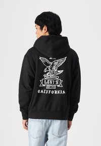 Felpa nera con cappuccio con una grafica bianca di un'aquila e testo del marchio sulla schiena. Materiale in cotone con vestibilità comoda e tasca a marsupio.
