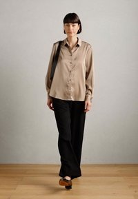 Beige selyem gombos ing, fekete széles szórású nadrág, barna és fekete cipők, modell egy fekete válltáskával, minimalista háttér.
