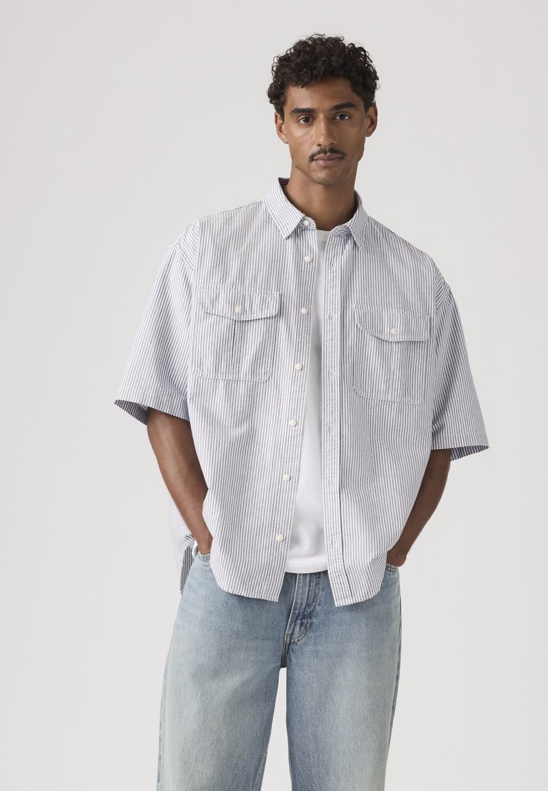 Jeune homme portant une chemise à manches courtes rayée bleu clair et blanc par-dessus un t-shirt blanc, avec un jean bleu clair, les mains dans les poches.