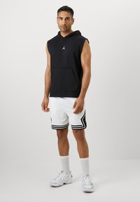 Svart hettejakke uten ermer med frontlomme, kombinert med hvite nettingshorts med svarte striper. Hvite trening-sko og sokker.