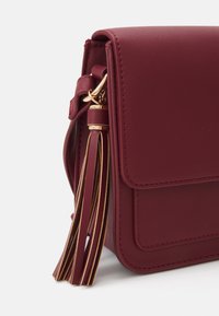 Borsa a tracolla in ecopelle bordeaux con chiusura a patta magnetica, con un dettaglio in metallo dorato e un lungo ciondolo a frange.