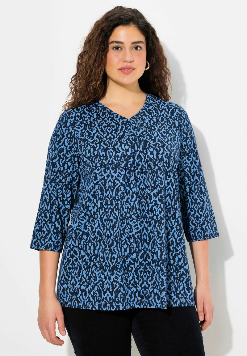 Modrý top s výstřihem do V, zdobený vzorovaným designem v tmavě a světle modré, s rukávy po tři čtvrtiny a volným střih. Textura látky je hladká.