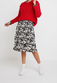 Röd stickad tröja med långa ärmar över en svart blommig mönstrad kjol. Vita sneakers kompletterar outfiten. Lagerdesign med kontrasterande mönster.