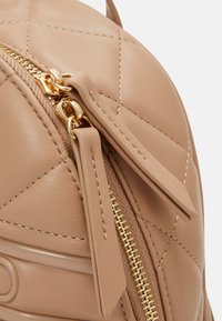 Tan quilted leather bag with a gold zipper, featuring dual pull tabs and smooth stitching details. Close-up view emphasizing texture and design.