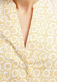 Blusa in tessuto bianco con motivo floreale giallo, dotata di placca frontale e dettaglio con un singolo bottone sul colletto. Tessuto morbido.