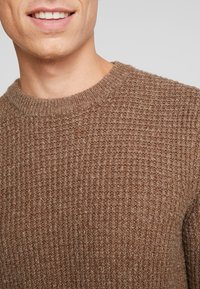 Nahbild av en person som bär en strukturerad brun stickad tröja med ribbad rundad halsringning, som visar den nedre delen av ansiktet och nacken.