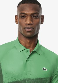 Grön polo shirt med krage, två knappar, gradientstruktur och en liten krokodil-logotyp på vänster bröst. Slät fäbrik.