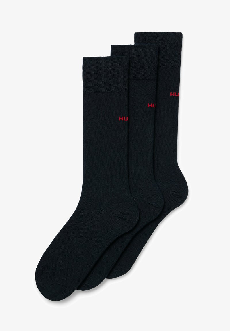 Chaussettes noires en coton dans un pack de trois, avec une bordure côtelée et un petit logo "HUGO" rouge sur le côté. Texture lisse et design classique.