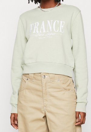 Persoon die een lichtgroene trui draagt met de tekst "FRANKRIJK Paris League" en beige broek met hoge taille, staand tegen een effen achtergrond.