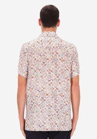 Uomo che indossa una camicia a maniche corte con un motivo floreale e geometrico colorato, di spalle, contro uno sfondo chiaro uniforme.