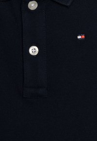 Jūras spēku polo krekls, kas izgatavots no teksturēta auduma, ar divu pogu apkakli, sudraba pogām un mazu izšūtu logotipu sarkanā, baltā un zilā krāsā.