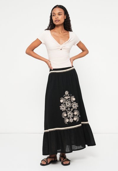 Črna vezenina maxi krila s poudarjeno robom, ki vsebuje cvetlične vzorce v krem in modri barvi. V kombinaciji z oprijetim svetlo rožnatim vrhom in sandali.