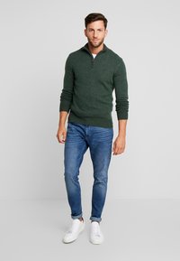 Suéter de punto verde oscuro con un cuello de media cremallera, llevado sobre una camiseta blanca, combinado con jeans de denim azules y zapatillas blancas.