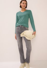 Grünes langärmliges V-Ausschnitt-Shirt, graue gerade Jeans und eine cremefarbene Umhängetasche. Die Schuhe sind hellbeige Sneakers. Minimalistisches Design mit klaren Linien.