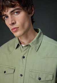 Light green button-up shirt with two chest pockets, black buttons, and a collar. Fabric texture appears durable with a slight weave pattern.