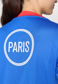 Mėlyna sportinė marškinė su raudonu apykakle, ant nugaros ryškiai vaizduojant balta apskritimą su tekstu "PARIS".