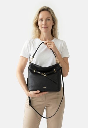 Žena v bílé košili a béžových kalhotách drží černou koženou kabelku se zlatým kováním, čelem k nám, na jednobarevném pozadí.