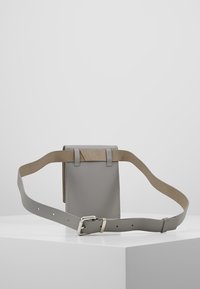 Šedá kožená taška přes rameno s hladkým povrchem, minimalistickým designem, nastavitelným popruhem a kontrastním hnědým akcentem na vrchu.