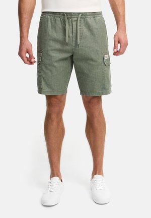 Mann iført grønne cargoshorts med snor i livet og sidelommer, samt hvite joggesko, stående mot en ensfarget bakgrunn.