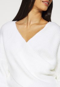 Weißer Strickpullover mit V-Ausschnitt, strukturiertem Stoff, weichem Finish und lockerem Schnitt, der entspannte Ärmel und einen drapierten Halsausschnitt betont.