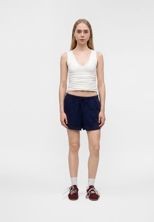 Ung kvinde står fremadvendt iført en hvid ærmeløs V-udskærings top, mørkeblå shorts med snøre, hvide sokker og mørkerøde sneakers.
