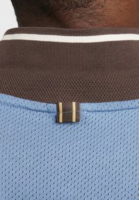 Blå nettingstoff t-skjorte med brun krage med hvite striper og en dekorativ brun etikett med to gyldne striper.