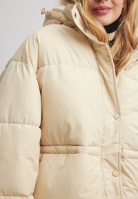 Béžová prešívaná puffová bunda s vysokým golierom, patentnými gombíkmi a nastaviteľnou kapucňou. Textúra je hladká a jemne lesklá.