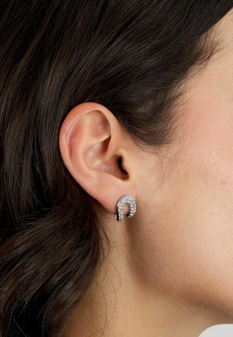 Orecchini in tonalità argento a forma di ferro di cavallo curvato, adornati con strass trasparenti, indossati sul lobo dell'orecchio con capelli scuri sullo sfondo.