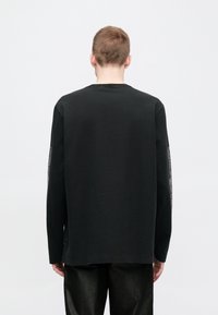 Juoda ilgomis rankovėmis medvilninė marškinėliai, turi apvalų kaklo iškirpimą ir grafinius raštus kairėje rankovėje. Lygus audinys, laisvas modelis.