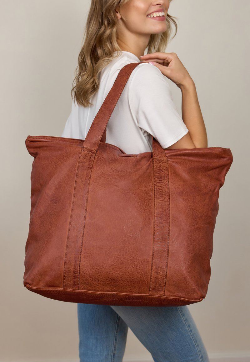 Rudos odos rankinė su minkštu paviršiumi ir dviem tvirtais diržais, kuri pasižymi matomomis siūlėmis ir erdviu dizainu.