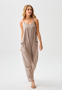 Hellbrauner Jumpsuit mit dünnen Trägern, seitlichen Taschen und entspanntem Schnitt. Aus weichem Stoff gefertigt, verfügt er über einen Rundhalsausschnitt und ausgestellte Beine.