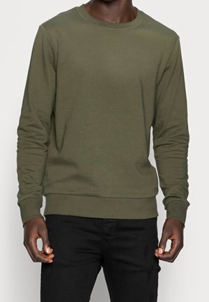 Homme portant un sweat-shirt kaki à manches longues avec col rond et un pantalon noir, debout devant un fond clair uni.