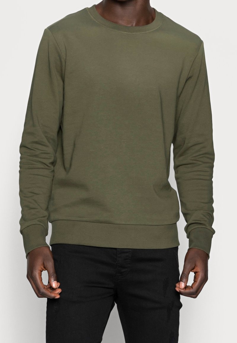 Homme portant un sweat-shirt kaki à manches longues avec col rond et un pantalon noir, debout devant un fond clair uni.