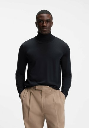 Mann trägt schwarzen Rollkragenpullover und beige plissierte Hose mit Händen in den Taschen, steht vor einem schlichten weißen Hintergrund.