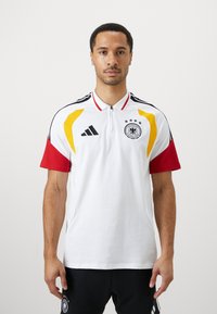 Hvit polo skjorte med røde og gule detaljer, krage med to knapper, tre stjerner over en sirkulær logo, svarte striper på skuldrene.