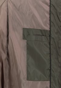 Binnenvoering van een jas met een rechthoekige olijfgroene stoffen zak genaaid op lichtbruin materiaal, met zichtbare stikseldetails.