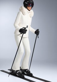 Costum de schi alb cu guler din blană falsă, mănuși negre și schiuri. Modelul poartă o cască neagră și ochelari de schi, ținând bețele de schi într-o poziție verticală.