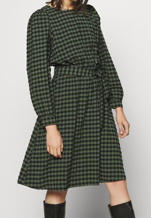 Vrouw draagt een groen-zwart geruit lange mouwen jurk met een bijpassende riem en zwarte kniehoge laarzen.