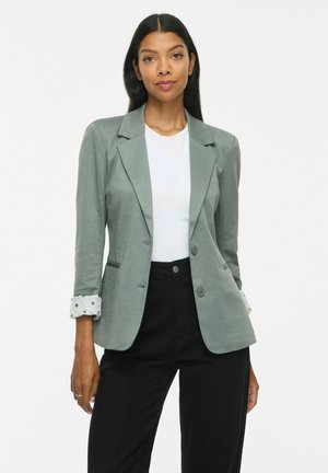 Femme aux longs cheveux foncés portant un blazer gris avec des poignets retroussés à pois, une chemise blanche et un pantalon noir, debout contre un fond uni.