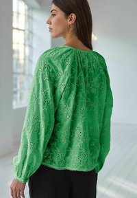 Blusa verde ricamata con maniche lunghe, motivi floreali, scollatura arricciata e delicato orlo scalloppato. Il tessuto appare leggero e testurizzato.