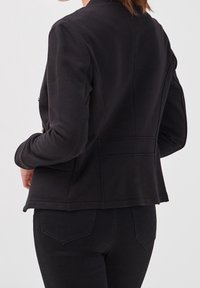 Woman wearing fitted black jacket and black jeans, shown from the back against a plain light background.