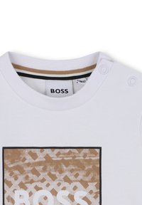 Weißes Baumwoll-T-Shirt mit Rundhalsausschnitt, mit braunem Grafikdruck in rechteckiger Form und zwei Druckknöpfen an der Schulter.