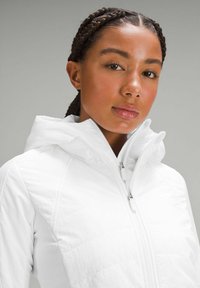 Veste blanche à fermeture éclair avec capuche, présentant un tissu texturé et un design matelassé. Détails de fermeture éclair visibles à l'avant.