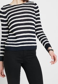 Sweter w paski w granatowym i białym kolorze, o dzianinowej fakturze, z długimi rękawami, ściągaczami przy mankietach i u dołu, w zestawie z czarnymi spodniami.