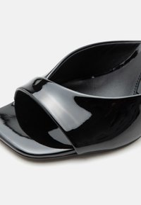 Γυαλιστερό μαύρο mule με ανοιχτά δάχτυλα, φαρδιά λουράκι και τετράγωνη μπροστινή όψη, απεικονιζόμενο από κοντινή πλευρική γωνία σε ανοιχτό φόντο.
