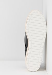 Chaussure synthétique noire avec une tige texturée et une semelle en caoutchouc blanche présentant des rainures horizontales. La chaussure est inclinée sur un fond blanc.