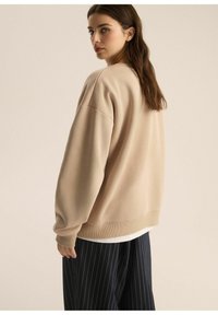 Beżowy, oversize'owy sweatshirt z prążkowanymi mankietami i dołem, zestawiony z granatowymi spodniami w prążki. Gładka tekstura, okrągły dekolt i opadające ramiona.