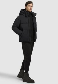 Sort pufferjakke med hætte, lynlåslukning og sidelommer, parret med sorte sweatpants og snørebånds kampstøvler.
