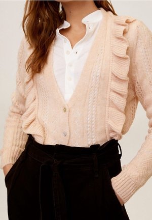 Gilet tricoté rose clair avec des bords volants, présentant un motif texturé et des boutons. Porté par-dessus une chemise blanche à col et un pantalon noir.