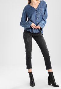 Femme portant une chemise en jean bleu boutonnée, un jean skinny noir déchiré avec des clous, et des bottines à talons noires, debout les mains jointes.